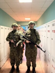 Dwóch uczniów klasy mundurowej w pełnym kamuflażu, hełmach i z atrapami broni stoi na korytarzu szkolnym między szafkami.