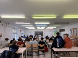 Grupa uczniów w sali lekcyjnej podczas prezentacji. Na środku znajduje się ekran interaktywny, a obok niego mapa Polski i biała tablica. Uczniowie siedzą w ławkach tyłem do obiektywu, słuchając nauczyciela.