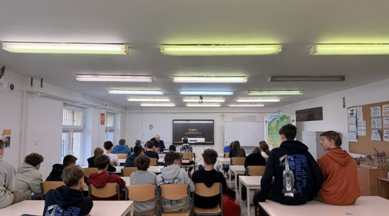 Grupa uczniów w sali lekcyjnej podczas prezentacji. Na środku znajduje się ekran interaktywny, a obok niego mapa Polski i biała tablica. Uczniowie siedzą w ławkach tyłem do obiektywu, słuchając nauczyciela.