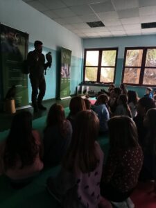 Mężczyzna w brązowym ubraniu sokolniczym stoi pośrodku sali, w lewej ręce trzymając na rękawicy ptaka drapieżnego, a prawą dłonią gestykuluje podczas prelekcji. Po obu stronach mężczyzny stoją dwa zielone roll-upy reklamowe Usługi Sokolnicze Rafał Przybylski, na których widać zdjęcia ptaków i logo. W tle po prawej stronie widać okno z widokiem na drzewa, a z przodu, na niskim, zielonym stojaku, siedzi jeszcze jeden ptak drapieżny. Sala ma biało-błękitne ściany i zieloną podłogę.