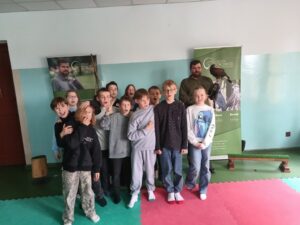 Grupa trzynastu dzieci w wieku szkolnym pozuje do zdjęcia pamiątkowego, stojąc w dwóch rzędach w sali lekcyjnej. Za nimi stoi mężczyzna w brązowym ubraniu sokolniczym, trzymając na rękawicy ptaka drapieżnego. Po lewej i prawej stronie grupy stoją dwa zielone roll-upy reklamowe Usługi Sokolnicze Rafał Przybylski. Dzieci są uśmiechnięte, niektóre pokazują znak pokoju, patrząc w obiektyw. Ściany są biało-błękitne, a podłoga zielona.