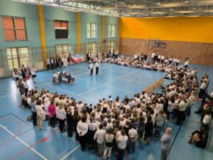 Szeroki półokrąg siedzących na parkiecie dzieci w strojach galowych (głównie białe koszule/bluzki i ciemne spodnie lub spódnice) zamyka przestrzeń z prawej strony i w głębi sali. Na pierwszym planie, tyłem do obiektywu, stoi zwarta grupa starszych uczniów oraz wychowawców obserwujących scenę. W lewej części kadru widoczny jest poczet sztandarowy ze sztandarem szkoły oraz opiekunowie